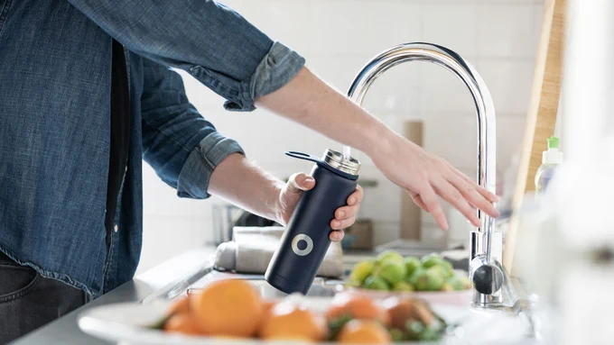 Person filling a bottle with water from a silver kitchen tap. Plates of fruit sit on the kitchen bench next to the sink.