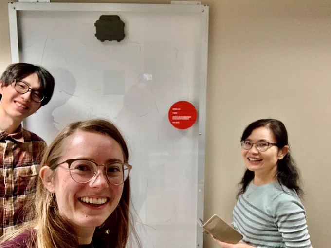 Three people pose for a photo in front of a cracked panel against a wall.