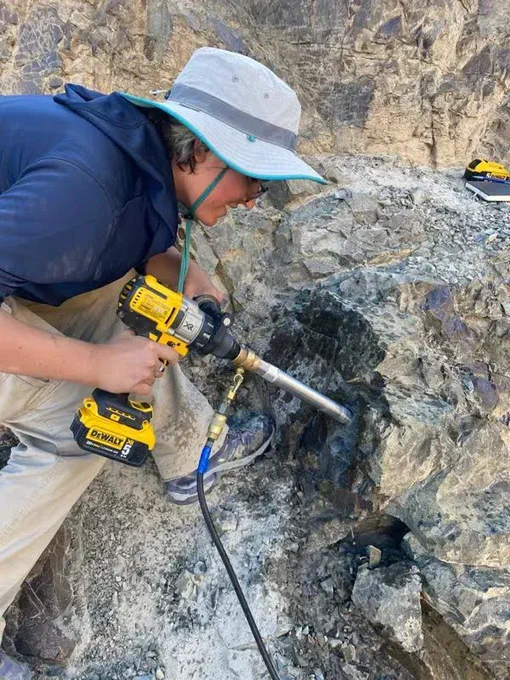 Kate Przydział, wearing a brimmed hat and holding a drill, drills into a large rock to collect a sample.