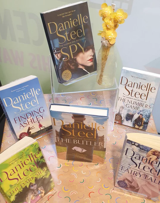 Six Danielle Steel books on a table including Fairytale, spy, the darkside, the numbers game, the Butler and Finding Ashley. There is also a vase with fake yellow flowers in it. 
