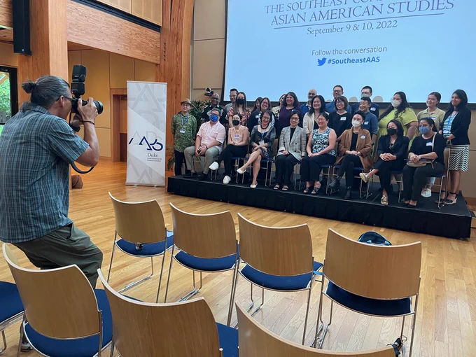 A photographer stands among chairs, pointing his camera toward 25 faculty, staff and postdocs assembled on stage next to an AADS Duke banner