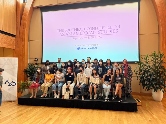 A group of more than 20 students pose seated and standing under a projected slide that says "The Southeast Conference on Asian American Studies - September 9 & 19, 2022"