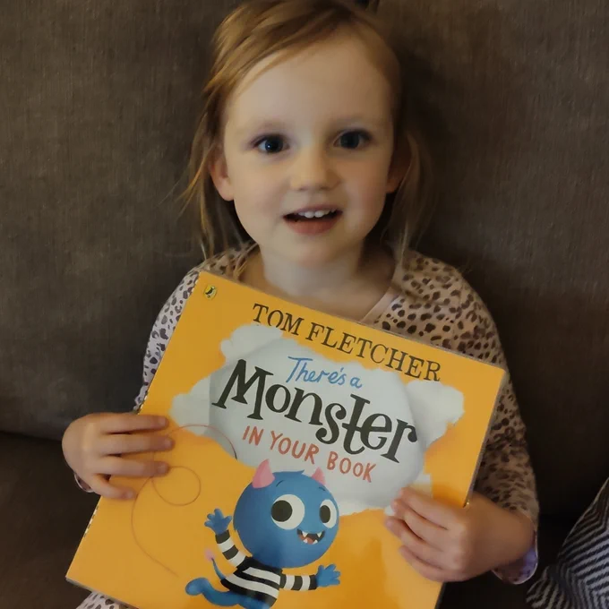 Photo of a little girl sitting on a brown sofa, holding a children's picture book. The book is called There's A Monster In Your Book and it is yellow and has an illustration of a small, smiling, blue creature with little horns and a tail.