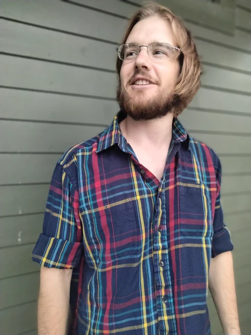 A photo of Matthew Hyslop standing against a green outside wall. He is wearing a blue plaid shirt, glasses, has a beard and medium-length brown hair.