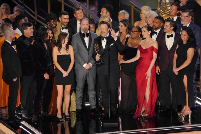 The cast of Ted Lasso stands together on the 2022 Emmys stage as Jason Sudeikis speaks into the microphone to accept the award for Outstanding Comedy Series.