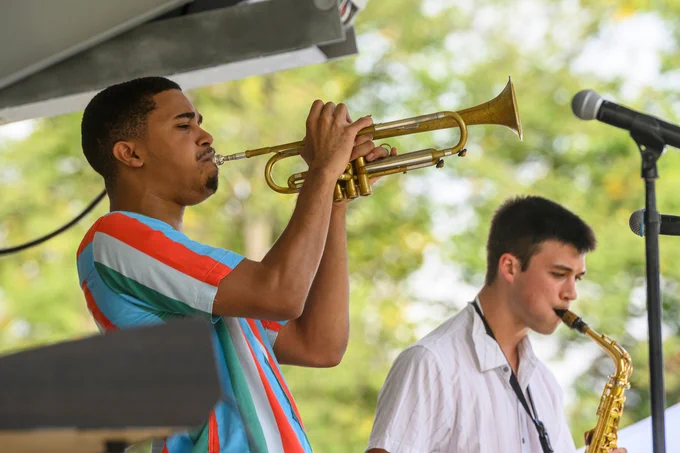 A trumpet player blows their instrument directly into a microphone, eyes closed. In the background, a saxophonist plays along.
