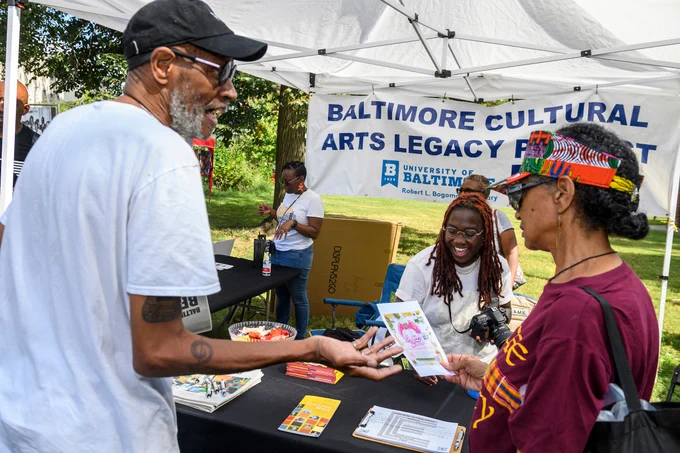 Two community members check out a booth promoting the Baltimore Cultural Art Legacy Project. Behind the booth, a representative for the project smiles.