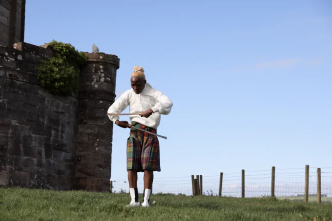 A film still from​‘Lagareh – The Last Born’. We see a Black womxn wearing a white sweater, a multi-coloured kilt and white and black boots. She is standing in a grassy field: her shoulders are raised and holding two machetes. In the background, there is an eighteenth-century folly and a simple fence with timber posts and metal netting. The sky is blue and there are no clouds.