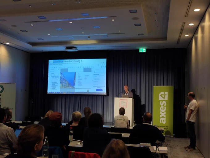 Vortragsraum des axes4 Day. Rufus Witt steht auf der Bühne und hält seinem Vortrag. Neben ihm wird seine Powerpoint auf einem Bildschirm angezeigt.