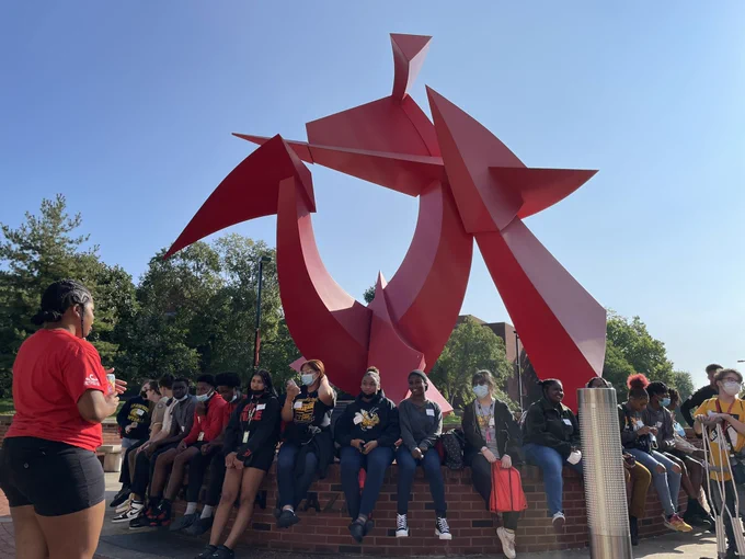 JCPS high school students sit near an art installation on the University of Louisville’s campus while a campus tour guide talks to the group.