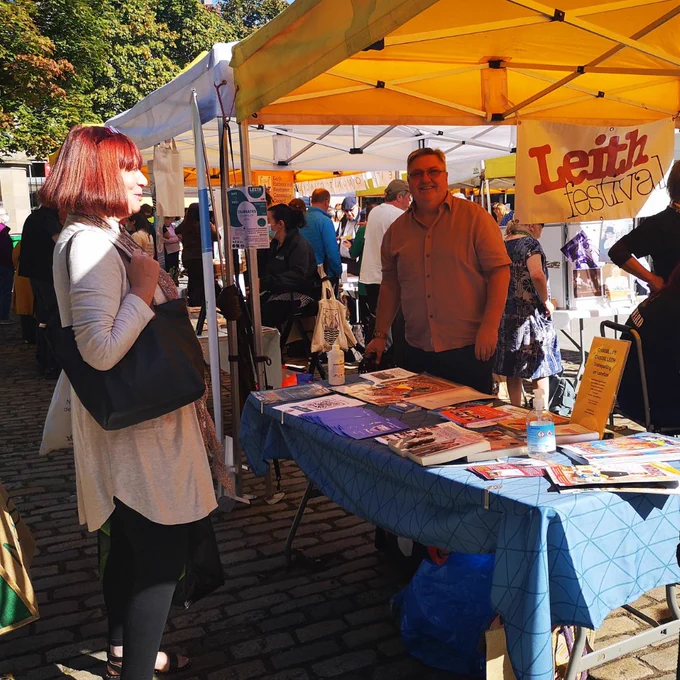 A woman with a white jacket and black handbag, with short red hair, talks to a man at a market stall under a bright yellow awning. The market stall has a sign that says Leith Festival.