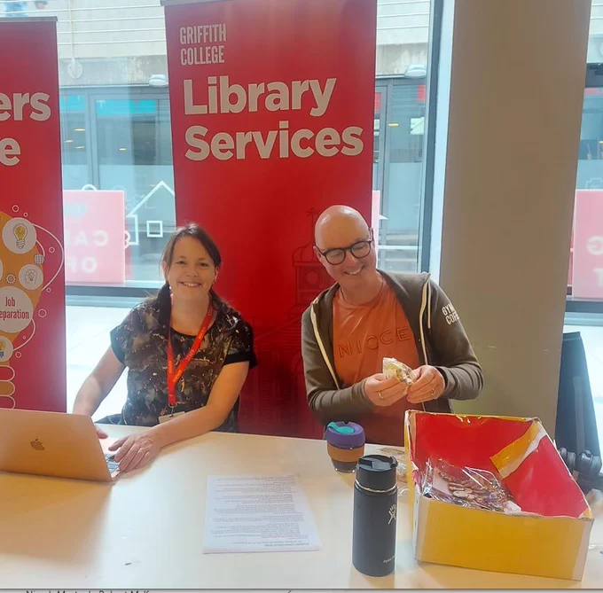 Griffith College librarians sitting at the Library Services desk at the 2022 induction. On the table in front of them is a box of badges.