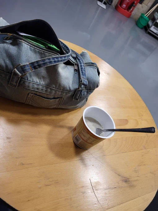 A slightly worn light blue denim handbag on a light wood circular table. The bag's zip is slightly open and a green USB cable cab be seen. The table is at Middlesbrough Town Hall. Next tother bag is an instant pot oat breakfast snack which the fabulous helpful staff located hot water and a fork so i could enjoy it. The fork is in the pot with the handle upright. In the background slightly out of focus stands a red fire extinguisher and a green mop bucket with two light brown mop handles visible. The floor around the table is a shiny grey colour suggesting it has been recently buffed. 
