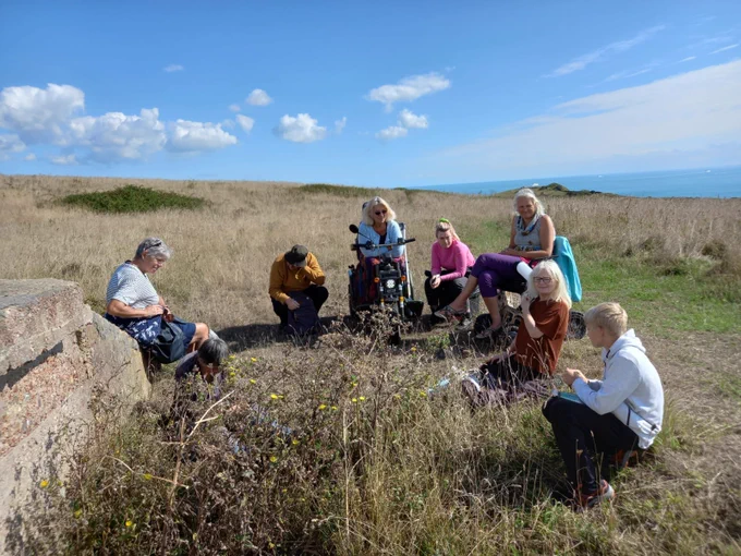 Several people gather to hear the story. Two women are in motor scooters. They are high on the Dover and Folkestone Heritage coast and the Channel is in the background.