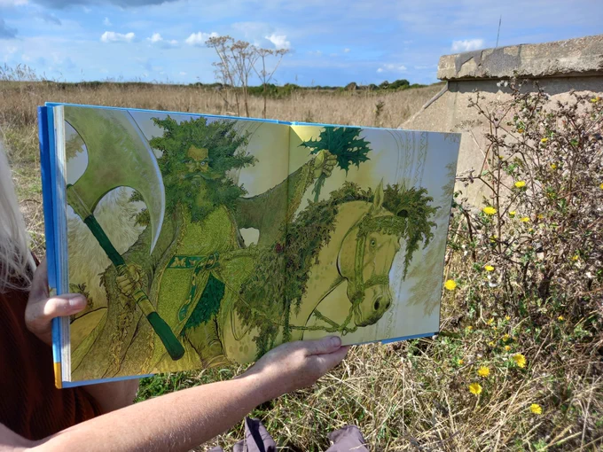 An illustration in the book of the Green Knight riding into Camelot on his green horse. He has a wild and bushy hair and beard and holds a bunch of Holly and a large axe. The background is grass and wildflowers.