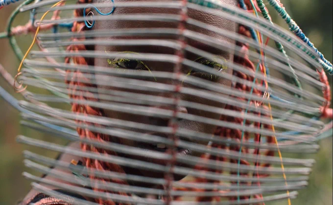 A Black person looks through an abstract helmet, that resembles a fence. They have orange and multicolored braids. They are outside, the background is out of focus, but lush. From Neptune Frost