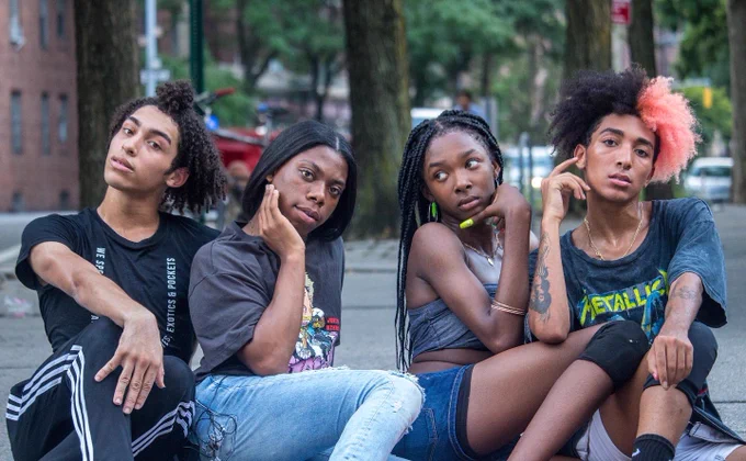 Four queer and trans Black young people sit on a curb in New York city. They wear dark colours, their hair is in four different styles, and they pose in a campy way for the camera. From Black As U R