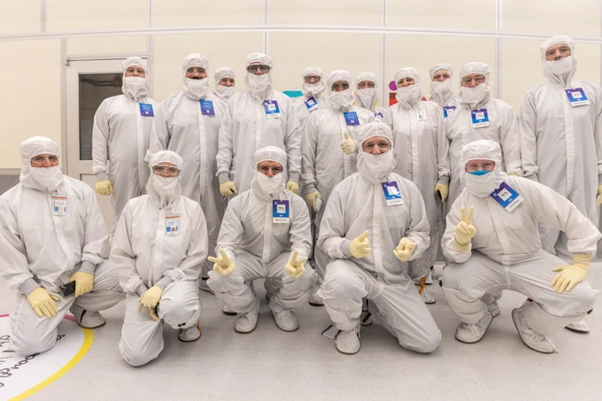 16 people pose for a photo wearing all white bunny suits, white gloves, blue ID badges and face and eye protection.