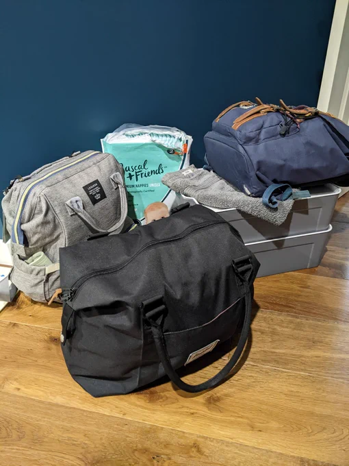2 backpacks, a weekend bag, 2 small storage bins, and a pack of nappies stacked together for a road trip.