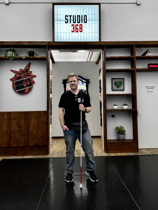 James stands holding his extended White Cane in front of walkway entrance into Studio 368. The front room features shelving that holds plants, art including wall art of a cartoon bomb exploding with a face. A light-up frame reads Studio 368 above the walkway. 