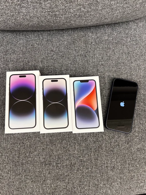 On a couch the boxes for iPhone 14 Pro Max, iPhone 14 Pro, and iPhone 14 are layed out. An iPhone 14 Pro is out of the box turning on with an Apple logo.