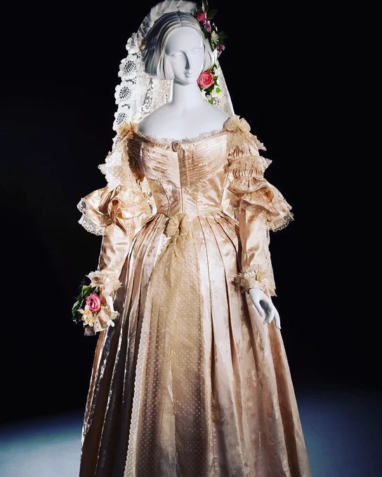 A cream satin wedding dress dating to 1838 with a pleated bodice, lace ruffles at the top of the sleeve and a white lace veil on top of the head