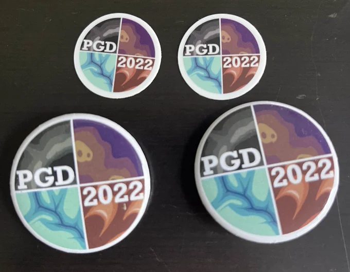 Circular buttons and smaller stickers featuring the 2022 division artwork - four quadrants in different colors showing elevation models of river valleys, sand dunes, mesas, and steep cliffs. PGD 2022 is written across the logo.