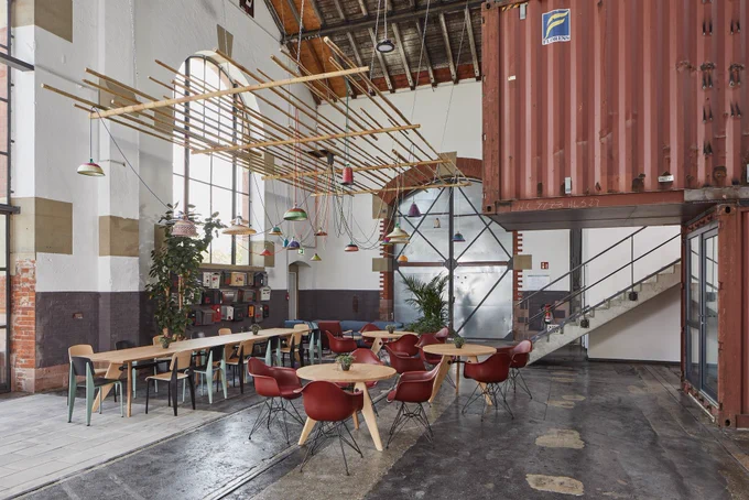 Eine alte Lokhalle neu als moderner Co-Working-Space umgebaut mit umgenutzten Containern, einer langen Tafel und Sitzgruppen mit Tischen; über ihnen hängen bunte Lampenschirme