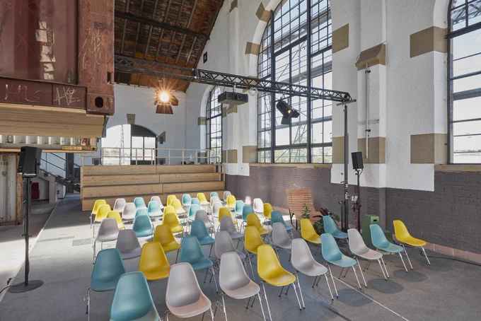 Eine alte Lokhalle neu als moderner Co-Working-Space umgebaut mit umgenutzten Containern, bunten Stühlen aus der Moderne sowie ein Treppenpodium