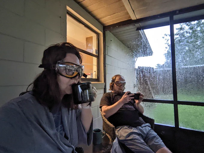 A man and woman having coffee in a screened in room in the rain. They are both wearing safety goggles. The woman has dark hair and is actively drinking from her cup. The man is looking at his phone.