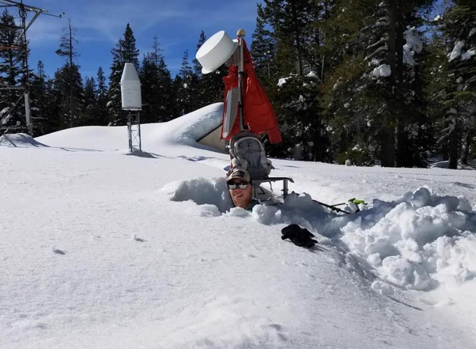 A person's head appears out of the top of a large snow pile. The snow is very deep and trees surround the area. Some equipment is seen in the background as well as a dark blue sky. 