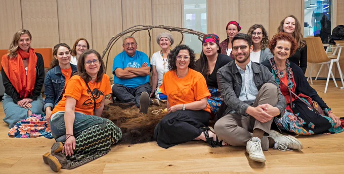 13 people sit on the floor to pose. Two are Elders sitting in the far back and the remainder are part of the art exhibit committee. 