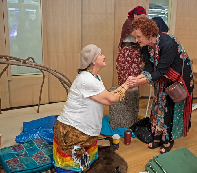 A woman on the right bends over to hold a woman (Elder)'s hands as a way to thank her. The Elder is on the floor sitting on her knees.
