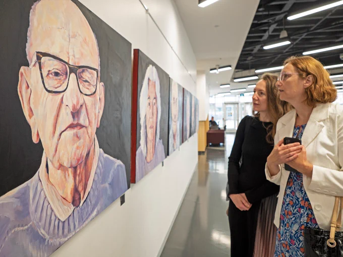 Two women look at paintings hanging on a wall.