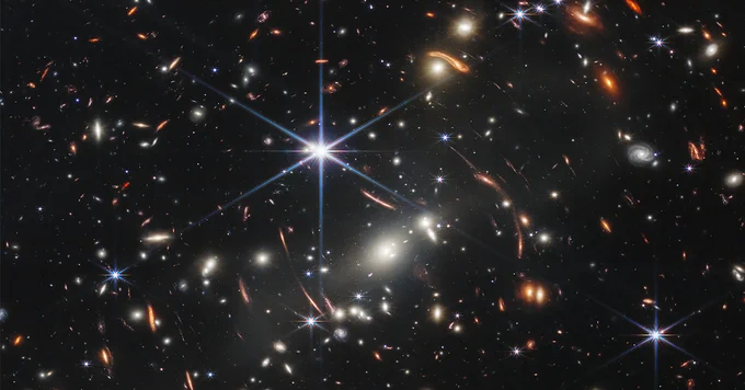 Image of Webb’s First Deep Field. Thousands of galaxies flood this near-infrared, high-resolution image of galaxy cluster SMACS. 3 stars in the centre shine very brightly against a black sky.
