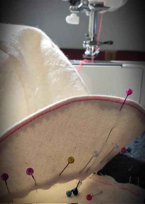 Many coloured pins in the brim of a off white cycle hat with hot pink stitches. White sewing machine threaded with pink thread in back ground 