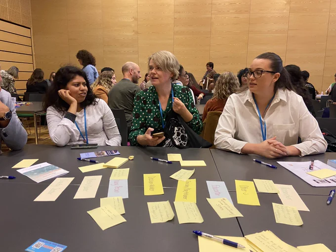 Three people sitting at a table full of sticky notes discussing ideas at an #iaymh2022 round table session.