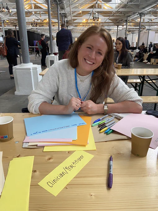 @drhollybear smiling at a table full of brightly coloured paper and sharpies, preparing for an #iaymh2022 session on implementation of evidence based practice