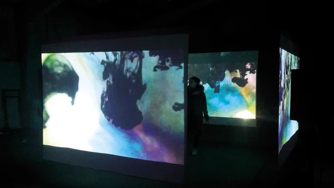Image of a person in a dark room surrounded by three screens. The screens are showing colourful artwork that depicts liquids and inks like substances