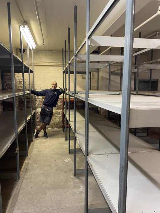 Matt leans against a rack of shelves, smiling with a thumbs up.