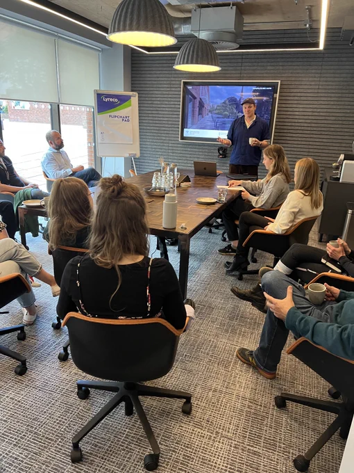 A group of professionals seated in a meeting room. A man stands at the front giving a talk in front of a digital screen.
