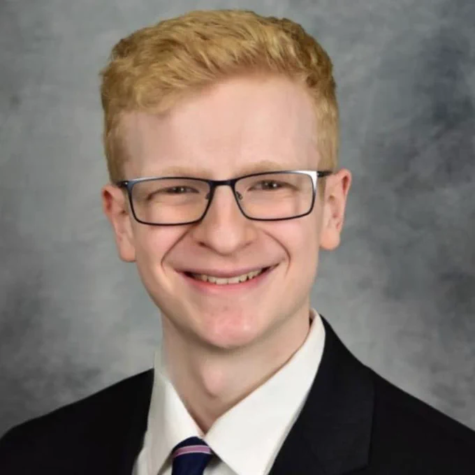 This photo shows Kevin standing in front of a grey backdrop. He is wearing a white collared shirt, a black suit jacket, a blue tie with a purple stripe, and glasses. Kevin has light blonde hair, and he is smiling at the camera.