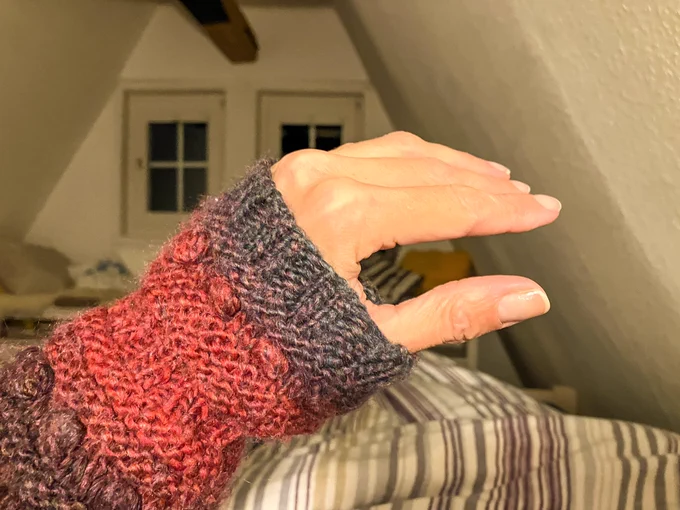 Foto meiner linken Hand mit lila-himbeer-graublauem Pulswärmer in Strukturstrick. Im Hintergrund ein gestreiftes Plümmo und eine spitze Giebelwand mit kleinen Sprossenfenstern.