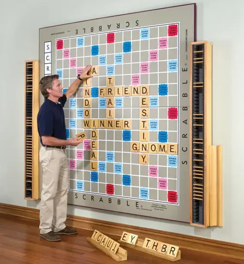 A man next to a scrabble board that is probably 10 feet tall attached to the wall.