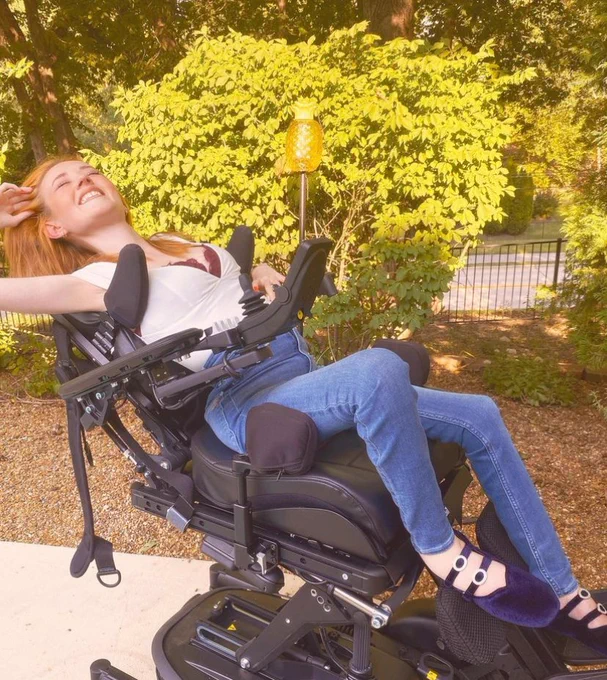 Madison, a white woman with red hair, in her wheelchair which is reclined back. She is smiling with her hand in her hair. Sh is wearing jeans, a white shirt, and blue velvet shoes.