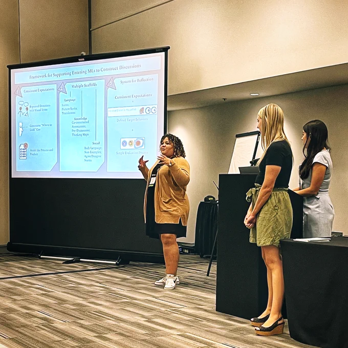 A photograph of Jenna Davis co-presenting a workshop before a large screen that displays the abridged text, "Framework for Supporting Entering MLs to Construct Discussions," while co-presenters Addison Barrack and Lindsay Roe stand to her side by a podium. 
