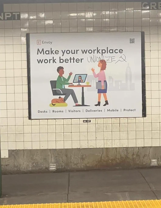 Train platform ad for company called Envoy with two office workers walking to each other and the word UNIONIZE written on it with black marker and a hammer and sickle 