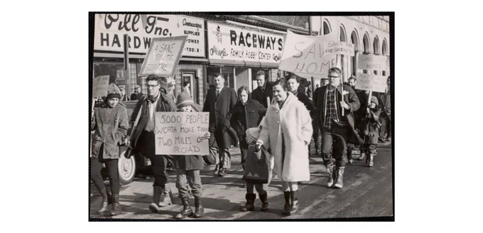 A photograph titled "Inner belt protest march in Cambridge" dated February 26th, 1966. The photograph shows protesters marching down a street. A child in the foreground is holding a sign that reads "5000 people are worth more than two miles of road."