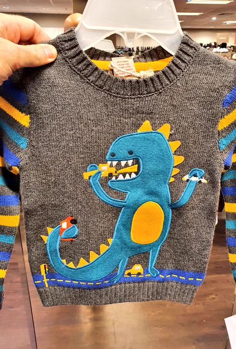 Auf einen grauen Strickpullover in Kindergröße ist eine riesige, Godzilla-artige blaue Echse gestickt, die auf einer Straße steht und Autos in die Höhe reißt und frisst