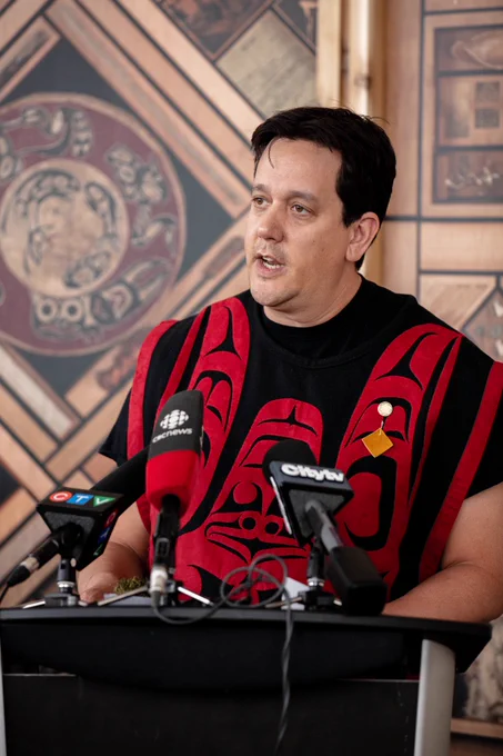 Artist Carey Newman stands at a podium with microphones in front of the Witness Blanket installation while wearing a red and black shawl with Coast Salish designs. Photo: CMHR, Keith Fraser.